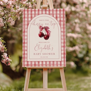 May include: A baby shower sign on an easel with a red and white gingham pattern. The sign reads "She's the cherry on top" with a cherry illustration. Below, it says "Welcome to Elizabeth's Baby Shower 04.24.XX".