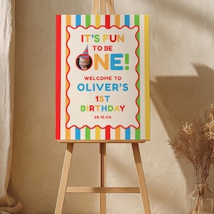 May include: A colourful birthday sign on a wooden easel. The sign reads "IT'S FUN TO BE ONE!" with the name "Oliver's" and "1st Birthday" displayed. The sign has a red, yellow, green, and blue striped border.