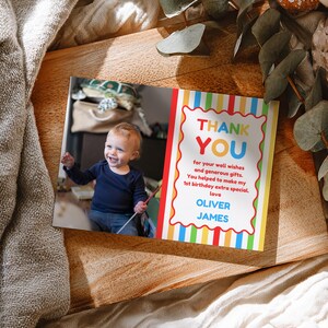 May include: A personalized thank you card with a photo of a smiling toddler. The card features colorful stripes and the words "THANK YOU" in large letters. The text reads "for your well wishes and generous gifts. You helped to make my 1st birthday extra special. love OLIVER JAMES."