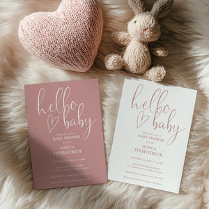 Op de afbeelding: Twee babyborrel uitnodigingen met de tekst "hello baby" in cursief, één roze en één wit. Een roze gebreid hart en een klein knuffelkonijn staan ook in de afbeelding. De uitnodigingen bevatten details voor de babyborrel van Jessica Fitzpatrick.