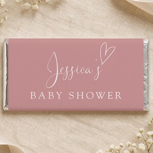 Può includere: Un involucro rosa per barretta di caramelle con la scritta "Jessica's BABY SHOWER" in corsivo bianco e un simbolo a forma di cuore. L'involucro è su uno sfondo di tessuto color neutro, con piccoli fiori bianchi.