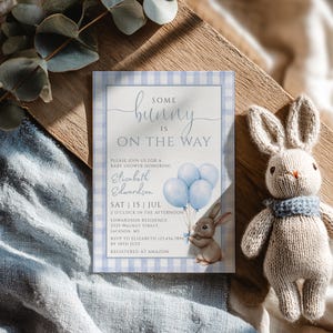 Könnte beinhalten: Eine Babyparty-Einladung mit blauem Vichy-Karo-Rand und dem Text "Some Bunny is on the Way". Die Einladung zeigt eine Aquarellillustration eines Hasen, der blaue Luftballons hält. Ein gestricktes Spielzeug-Häschen mit blauem Schal liegt daneben.