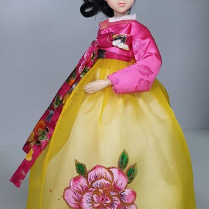 May include: A doll wearing a traditional Korean Hanbok dress. The dress is yellow with a large pink floral design. The top is pink with a decorative sash. The doll has black hair and is posed against a neutral background.