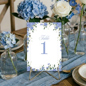 May include: A white table number card with the word "Table" and the number "1" in light blue. The card is decorated with blue floral accents and sits on a gold stand. Blue hydrangeas and white roses are in glass vases.