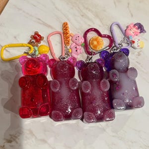 May include: Four colourful gummy bear keychains in red, pink, and purple. Each bear has a different colour and glitter effect. The keychains also feature various charms, including a bear, a heart, and a small figure.