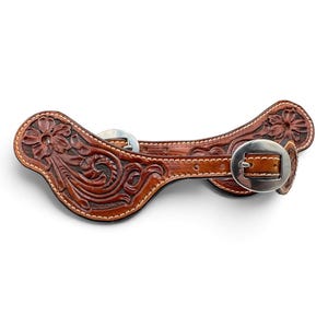 May include: A brown leather spur strap featuring intricate floral and scroll tooling patterns. The strap includes a polished silver-tone metal buckle and a matching metal keeper, with visible light-colored stitching along the edges of the leather.
