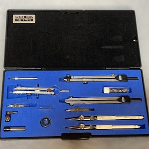 May include: A black case with a blue interior holds a set of drafting tools. The tools include compasses, dividers, and other drawing instruments. The case has a label that reads "UCHIDA KD-TYPE".