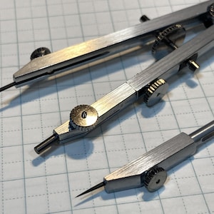 Può includere: Tre compassi da disegno color argento e un piccolo strumento divisore sono disposti su una superficie a griglia. I compassi hanno punte affilate e rotelle regolabili per misurazioni precise. Gli strumenti sono progettati per il disegno tecnico e la geometria.