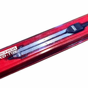 Può includere: Un compasso color argento, etichettato "UCHIDA KD-TYPE", è racchiuso in un astuccio di plastica rosso. Il compasso ha un'impugnatura nera e una punta. L'astuccio è rettangolare e ha un coperchio trasparente.