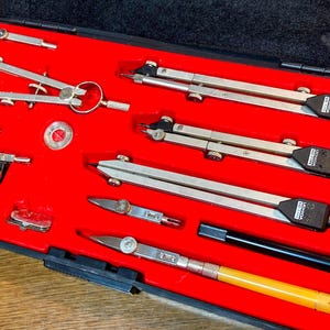 Puede incluir: Un juego de dibujo vintage en un estuche negro con interior rojo. El juego incluye varios compases, divisores y otras herramientas de dibujo de color plateado. Las herramientas están cuidadosamente dispuestas, mostrando su precisión y artesanía.