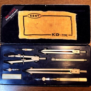 May include: Open black case containing a set of drawing instruments. The set includes compasses, dividers, and other tools, with a yellow cloth and the text "KENT" and "KD-TYPE". The case is open on a wooden surface.