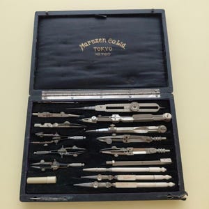 May include: An open, vintage drafting set in a black case. The case is lined with black velvet and contains various silver-toned drawing instruments. The lid has gold lettering that reads "Maruzen Co. Ltd. TOKYO No. 760".