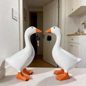 May include: Two white goose figurines with orange beaks and feet, each holding a key in its beak. The geese are standing on a white surface, with a neutral background. The figurines are a decorative item.