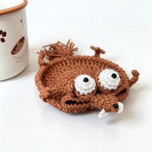 May include: A brown crocheted coaster shaped like a cartoon character, featuring large white eyes, a white snout, and a brown tail. The coaster rests on a white surface, with a white mug with brown details in the background.