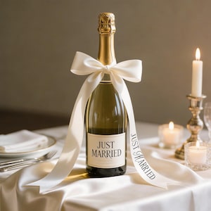May include: A champagne bottle with a gold foil top and a white ribbon tied around it. The bottle has a label that reads "JUST MARRIED". The ribbon drapes down the front of the bottle and has the words "JUST MARRIED" printed on it. Candles and a place setting are in the background.