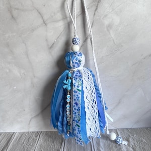May include: A blue and white tassel ornament with a beaded accent spelling "BE STILL". The tassel features blue ribbons, lace, and floral fabric. A white cord is attached for hanging, and the ornament includes decorative beads.