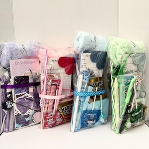 May include: Four clear cellophane gift bags filled with various items. Each bag is tied with a ribbon and contains items like pens, snacks, and small notebooks. One bag has a purple theme, another pink, a third blue, and the last green.