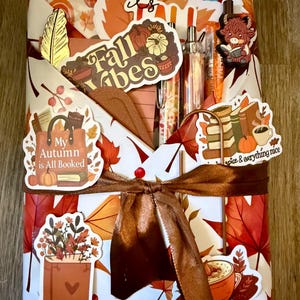 May include: A rectangular gift package wrapped in white paper with a pattern of orange and red autumn leaves, tied with a brown satin ribbon. It is decorated with various stickers featuring pumpkins, books, coffee mugs, and the text Fall Vibes.