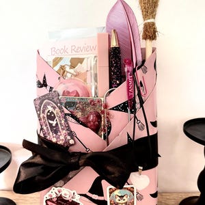 May include: A pink gift box with a black bow, witch hat and broomstick pattern, filled with various items. Includes a feather, a small broom, a pen, a lipstick, a book review, and stickers. The stickers feature a stack of books and a cat.