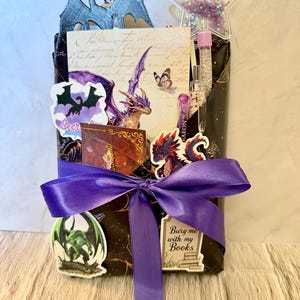 May include: A gift-wrapped package in black paper with a purple satin ribbon bow, decorated with dragon-themed stickers, a star-shaped shaker topper, and a card featuring a purple dragon. A small sign reads, Bury me with my Books.