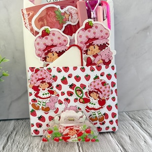May include: A collection of Strawberry Shortcake-themed items. Includes a white envelope with stickers, a pink pen, a pink diamond-shaped pen, and a clear hair clip with a Strawberry Shortcake design. The envelope is decorated with Strawberry Shortcake illustrations and strawberries.