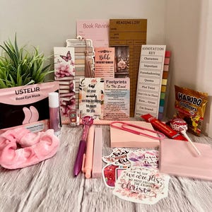 May include: A collection of pink and white stationery and self-care items, including a rose eye mask, pens, stickers, a notepad, and a bag of gummy bears. There are also several cards with text.