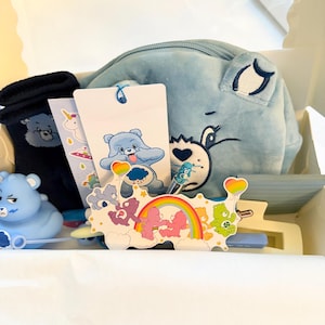 May include: A gift box containing a light blue plush pouch with a bear face, a pair of dark blue socks, a blue bear-shaped figurine, a white bookmark with a blue bear illustration, a sticker sheet with colorful bears and a rainbow, and a pen.