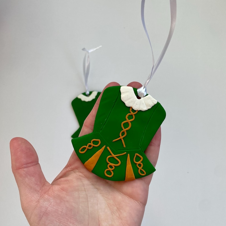 May include: Green ornament in the shape of an Irish dance dress, featuring gold detailing and a white collar. The ornament hangs from a white ribbon. Another ornament is visible in the background.