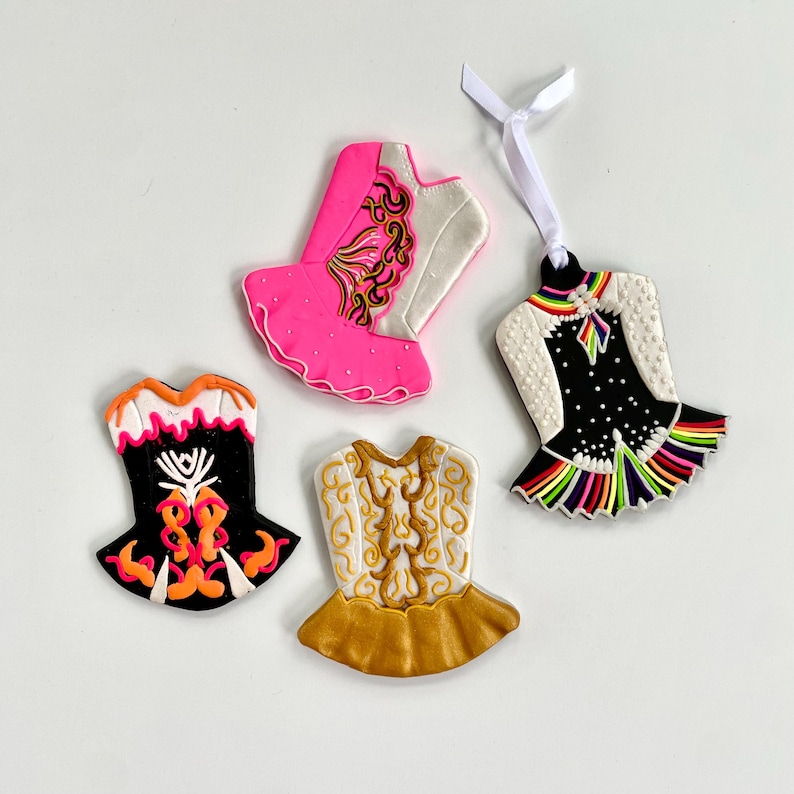 May include: Four colorful Irish dance dress ornaments. One is black with orange and white accents, another is gold and white, a third is pink and white, and the last is black with rainbow trim and a white ribbon.
