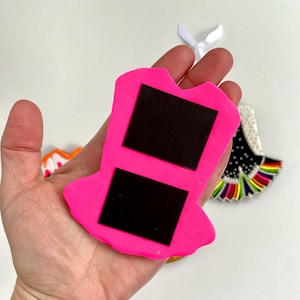 May include: A bright pink magnet shaped like a dress, with two black rectangular magnets on the back. The magnet is held in a hand, with other decorative magnets in the background.