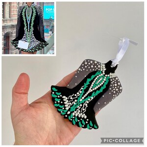 May include: A black and gray ornament shaped like an Irish dance dress, with white and teal accents. The ornament has a white ribbon for hanging. The design is a miniature replica of an Irish dance dress.