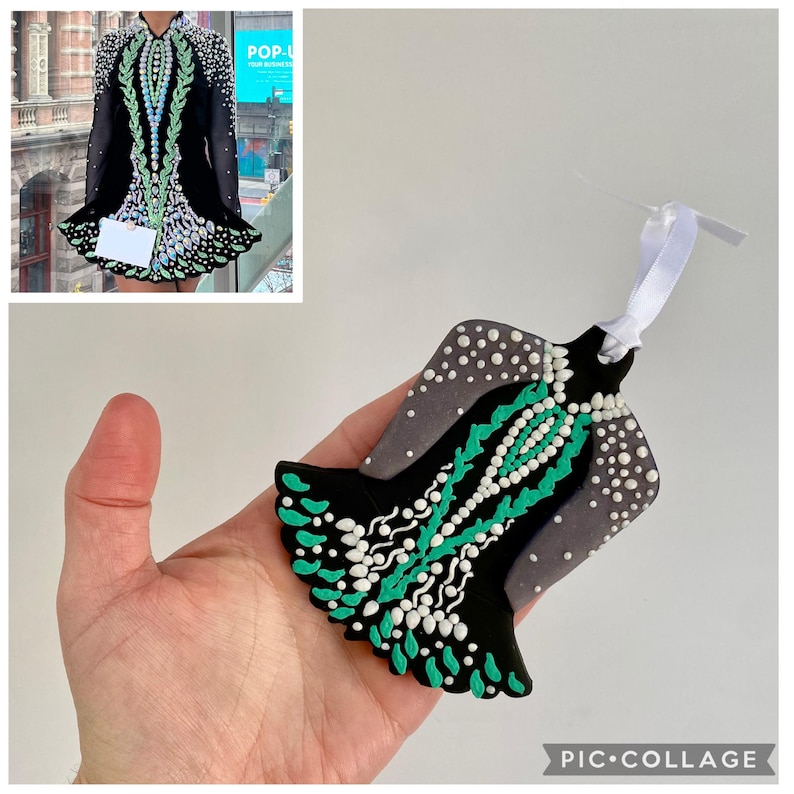 May include: A black and gray ornament shaped like an Irish dance dress, with white and teal accents. The ornament has a white ribbon for hanging. The design is a miniature replica of an Irish dance dress.
