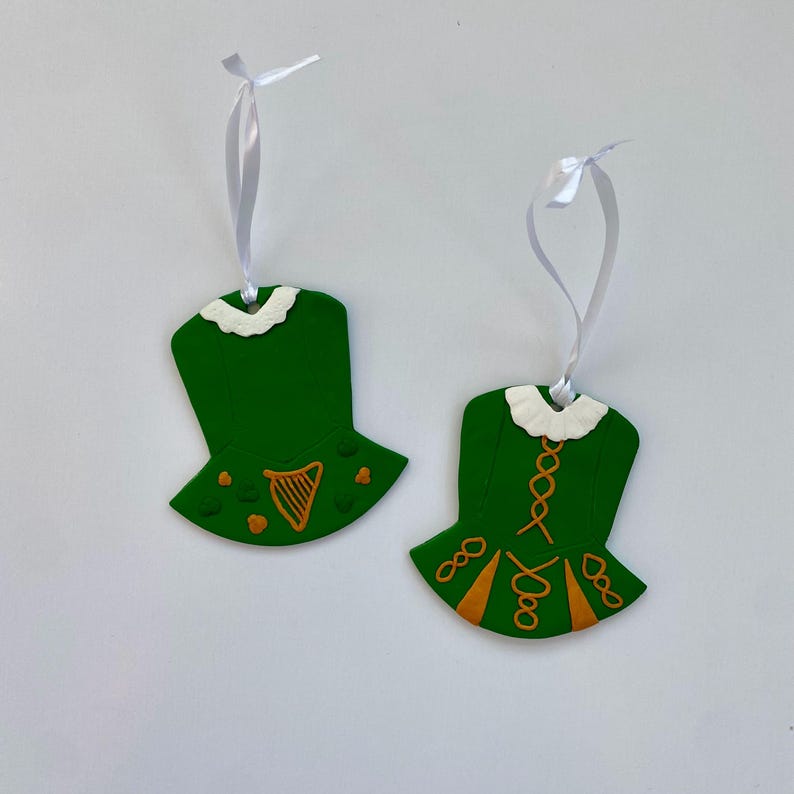 May include: Two green Irish dance dress decorations with white ribbon hangers. The dresses feature white collars and orange detailing, including harps and decorative patterns. The ornaments are set against a white backdrop.