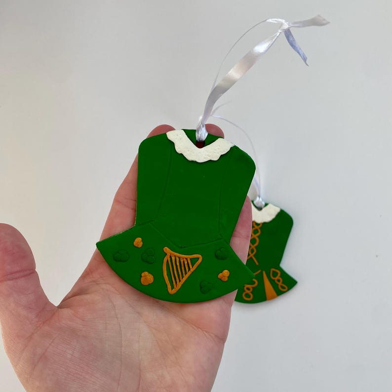 May include: Two green wooden ornaments in the shape of Irish dance dresses. Each has a white collar, orange harp and shamrock details, and a white ribbon for hanging. The ornaments are held in a hand.