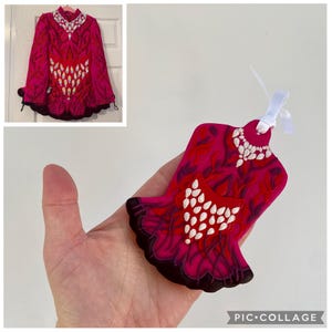 May include: A vibrant fuchsia-colored ornament shaped like a dress, featuring white bead-like accents and a dark brown scalloped hem. The design includes a heart-shaped detail with white embellishments. A matching dress is hanging in the background.