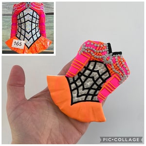 May include: A vibrant, decorative cookie shaped like a dance costume. The cookie features a bright pink bodice with silver bead accents, a black and white geometric design, and an orange skirt. The number "165" is visible on a tag.