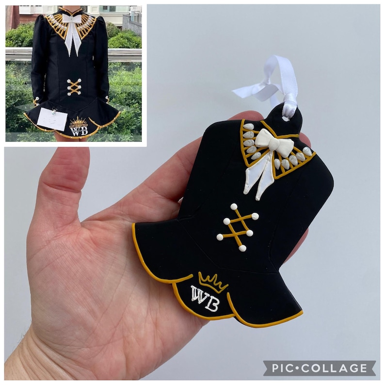 May include: Black ornament shaped like a traditional Irish dance dress. The ornament has gold trim, white accents, and a white bow. The initials "WB" are displayed on the bottom of the ornament. A white ribbon is attached for hanging.