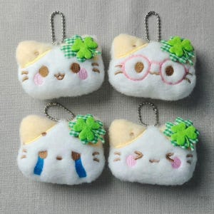 May include: Four white plush cat-shaped keychains with yellow accents. Each keychain has a different facial expression and a green clover accent. One cat wears pink glasses. Each keychain has a silver metal loop for attachment.