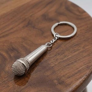 May include: A silver-colored microphone keychain with a mesh head and a ring for keys. The keychain is made of metal and is designed to resemble a miniature microphone. The keychain is on a wooden surface.