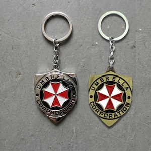 May include: Two keyrings with the Umbrella Corporation logo. One is silver-coloured, the other is gold-coloured. Each has a shield-shaped pendant with a red and white umbrella design and the words "Umbrella Corporation".