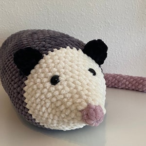May include: A crocheted plush toy shaped like an opossum. The toy is gray, white, and pink. It has black eyes and a pink nose.