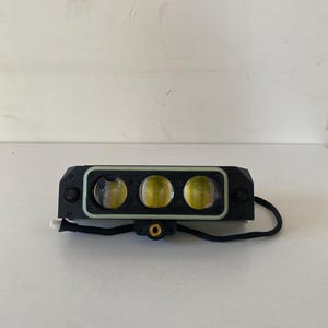 May include: A rectangular black LED light bar featuring three circular lenses with yellow-tinted glass. The housing has a light-colored border around the lenses, two side-mounted buttons, and a central mounting point with a gold-colored threaded insert.