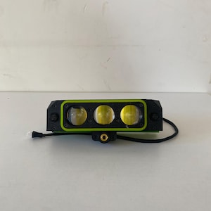 May include: A rectangular black LED light bar featuring three circular lenses with yellow-tinted glass. The housing is accented with a bright lime green border and includes a central mounting point with a gold-colored threaded insert and a black cable.