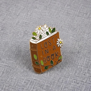 May include: A brown enamel pin shaped like a book with the text LOST IN A BOOK written in black. The book features white daisy flowers and green leaves on the cover and pages. The design includes gold-coloured metal outlines and details.