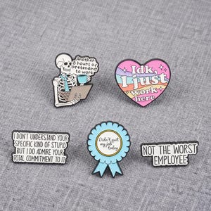 May include: Five enamel pins featuring workplace humour. One shows a skeleton with a laptop and coffee, one is a heart with rainbow stripes, one is a blue ribbon award, and two are rectangular pins with black text on white backgrounds.