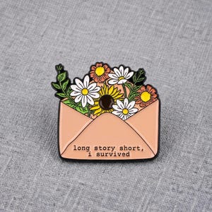 Op de afbeelding: Een perzikkleurige emaille speld in de vorm van een envelop die overloopt van diverse bloemen, waaronder witte margrieten en gele zonnebloemen. De tekst "long story short, i survived" staat in het zwart op de voorzijde van de envelop.