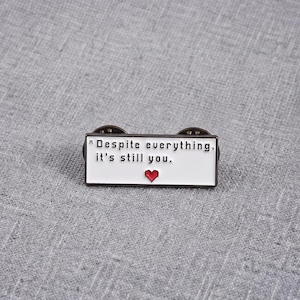 Puede incluir: Un pin de esmalte rectangular con fondo blanco y un borde negro fino. El pin presenta un texto negro pixelado que dice "* Despite everything, it's still you." y un pequeño icono de corazón rojo pixelado debajo del texto.