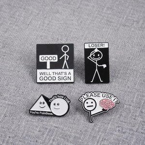 Op de afbeelding: Vier emaille pins met zwart-witte ontwerpen. Eén vierkante pin toont een stokfiguur met de tekst 'WELL THAT'S A GOOD SIGN'. Een rechthoekige pin zegt 'LOSER!', een driehoekige pin 'You're Pointless.' en een pin met een afbeelding van een brein zegt 'PLEASE USE IT'.