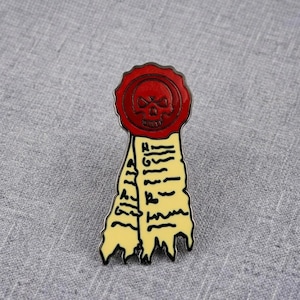 Peut inclure: Une broche en émail métallique en forme de sceau rouge circulaire avec une icône de crâne sur le dessus, attachée à deux rubans couleur crème qui pendent. Les rubans présentent un texte noir écrit à la main sur leur surface.