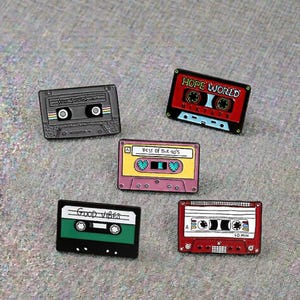 May include: Five cassette tape enamel pins in different colours. The pins feature text including "Good Vibes" and "Hope World Mixtape". The pins are black, red, green, pink, and yellow.